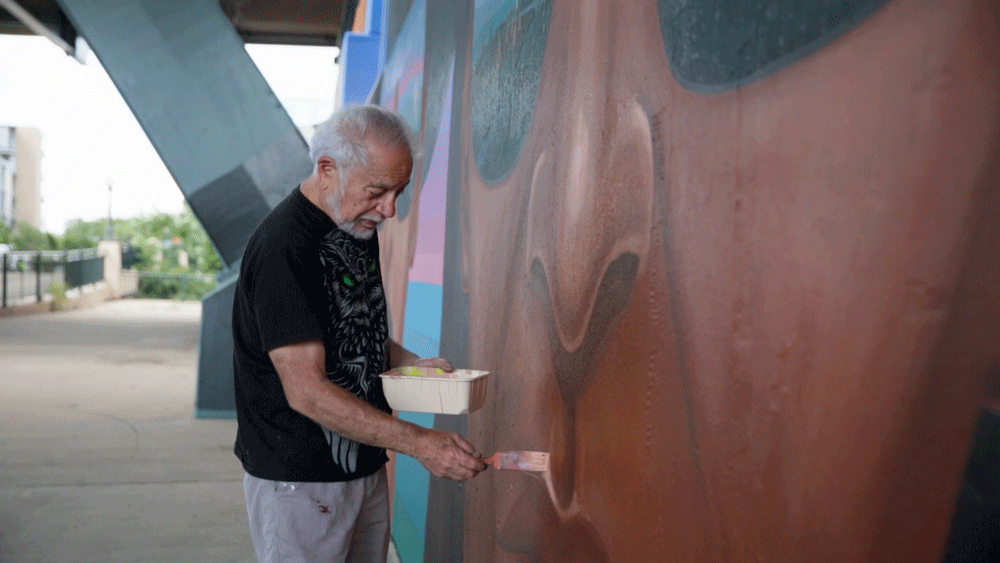 Emanuel Martinez adds tan paint to the upper lip of a large, dark-skinned face. He is wearing a black T-shirt and grey shorts.