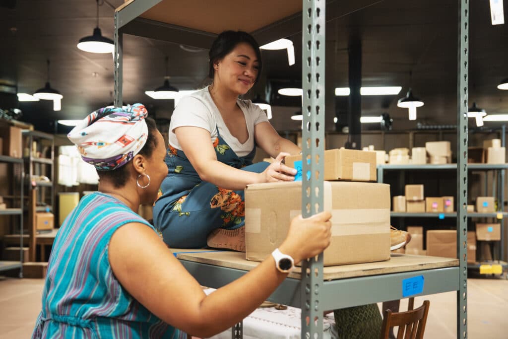Two women are in a room full of cardboard boxes. One sitds on a shelf with two boxes in front of her, while the other owman stands, back to the camera, looking at the women sitting on the shelf.