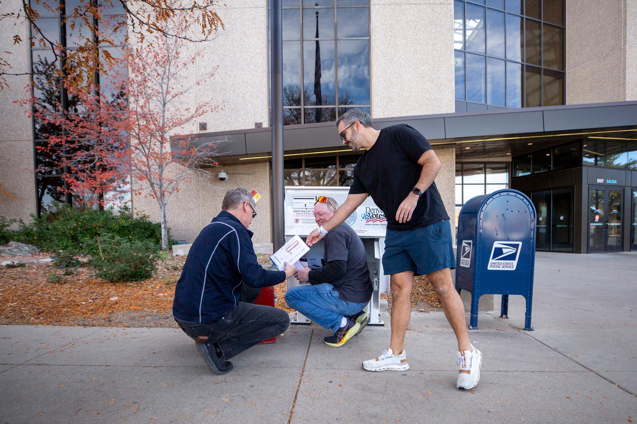 Three men are outside a building, one standing and two kneeling near a ballot drop box. The standing man, dressed in a black t-shirt, blue shorts, and white sneakers, is handing his ballot to the kneeling men, who are both election workers.