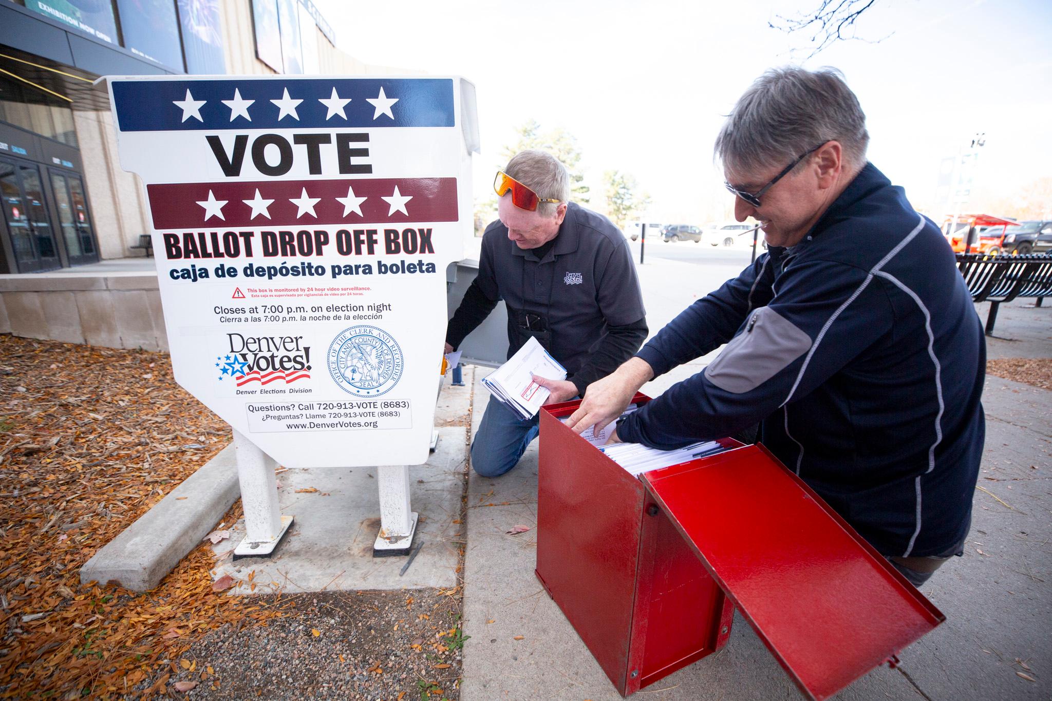 Two men, both election workers kneel next to a ballot box to collect ballots before stuffing them in a secure red metal box.