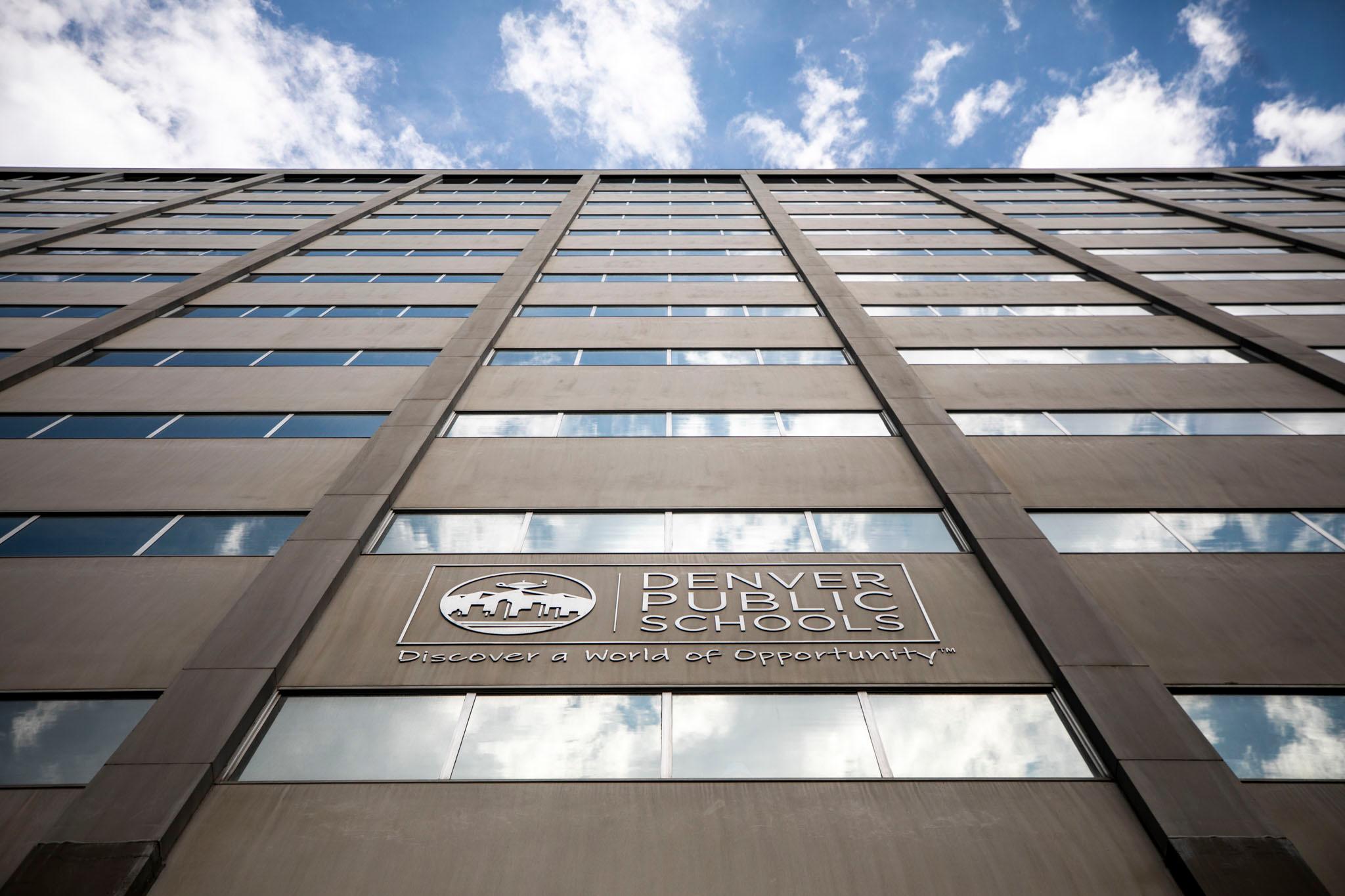 A building stretches into a blue sky, windows reflecting clouds above, with a sign on the side that reads "Denver Public Schools."