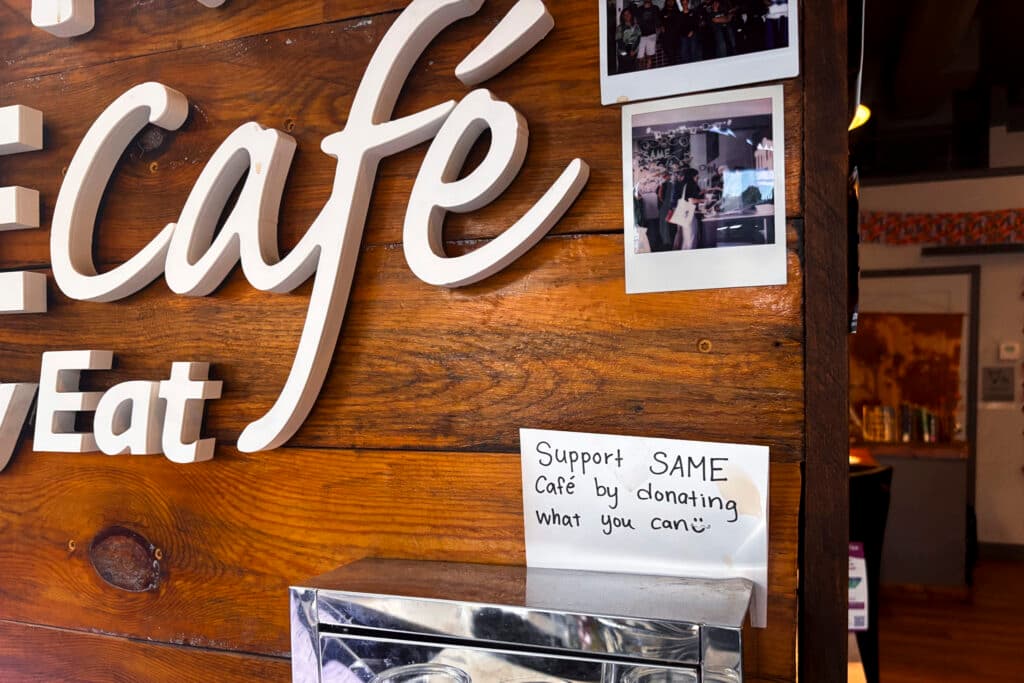 A sign above a donation box against a reclaimed wood wall that reads, "Support SAME cafe by donating what you can " with a smiley face.