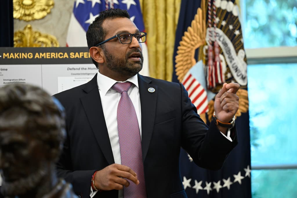 A man in a suit and glasses gives a speech in front of flags and a poster board that partially reads "Making America Safer."