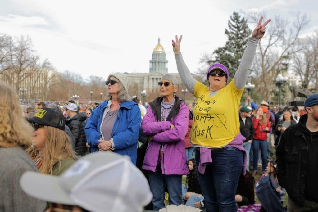 Three women stand in the crowd, wearing colorful light jackets and beanies. One woman's yellow shirt reads "Impeach Trump Deport Musk."