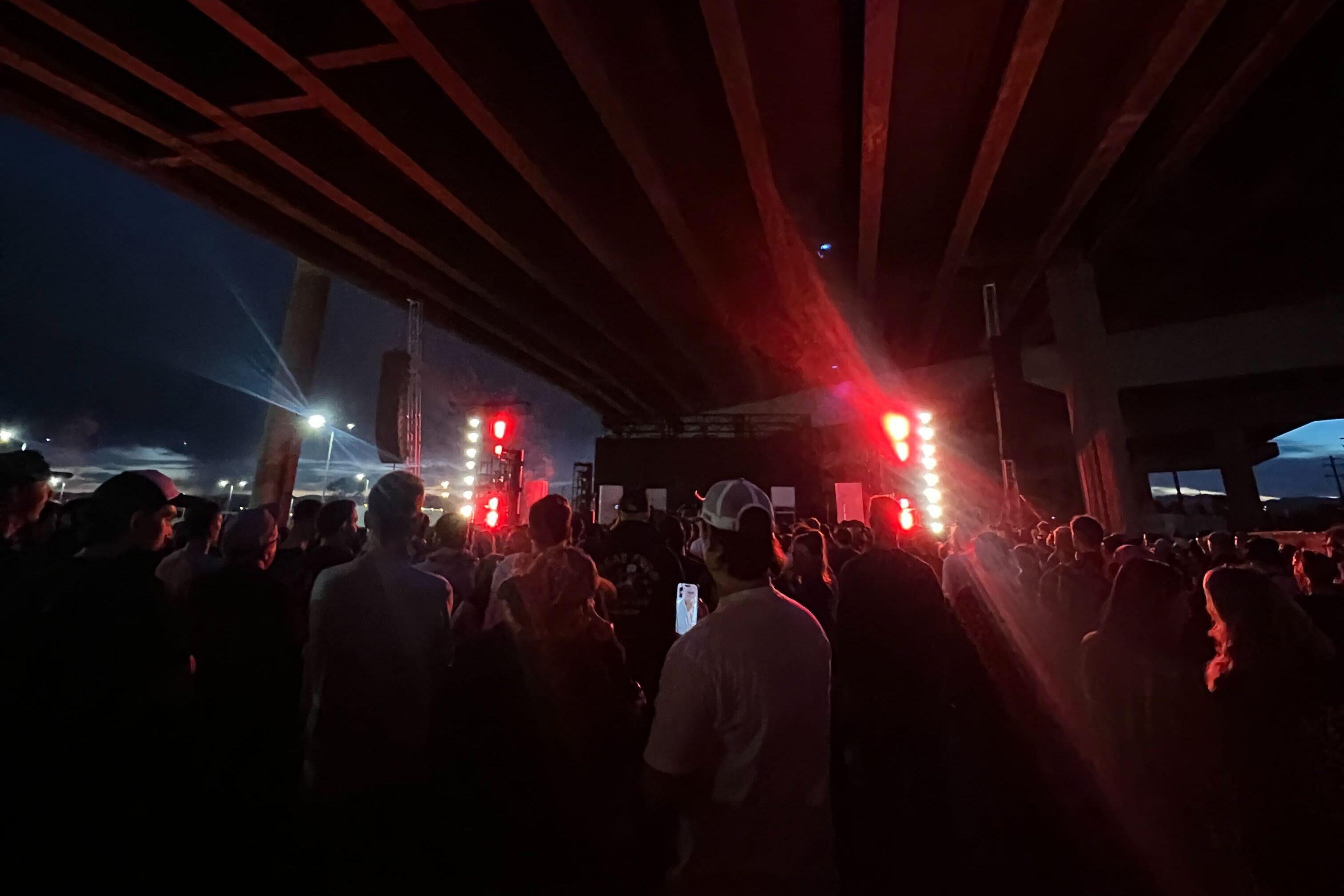 A large group of people are gathered under a highway, watching a performance with flashing red and white lights. The audience is standing close to each other, and some are holding cell phones.
