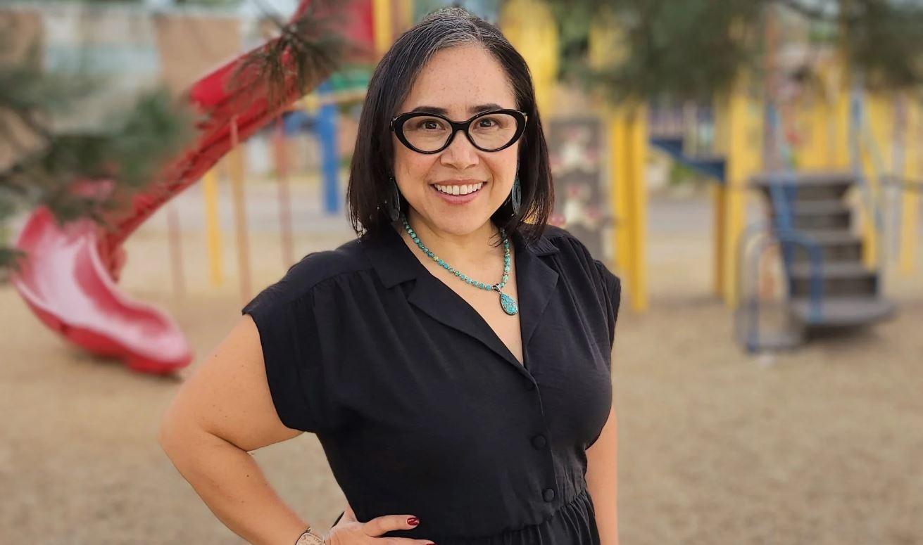 A woman wearing glasses, a turquoise necklace and a black dress smiles while standing outdoors in front of a playground with a red slide and yellow climbing equipment.